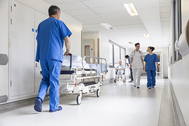 Eine Krankenschwester, die ein Bett in einem Krankenhausflur hinunterrollt. Enterprise Asset Management (EAM) wird in Gesundheitseinrichtungen eingesetzt, um die Einhaltung von Vorschriften zu unterstützen.