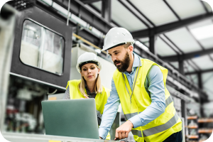 Man and woman in white hardhats and high-visibility vests looking at a laptop in a warehouse